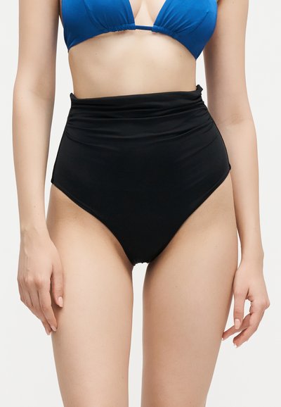 Femme portant un haut de bikini bleu et un bas de bikini noir taille haute sur un fond clair uni.
