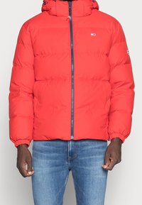 Rote Daunenjacke mit Kapuze, Reißverschluss und elastischen Bündchen. Besitzt einen navyblauen Akzent-Reißverschluss und ein kleines Logo auf der linken Brust.