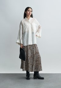 Γυναίκα που φοράει διαφανές λευκό πουκάμισο με κουμπιά, φούστα με λεοπάρ σχέδιο, μαύρες μπότες με μεταλλικές αλυσίδες, κρατώντας μαύρο φάκελο σε γκρι φόντο.