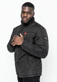 Svart vadderad jacka med hög krage, röd logodetalj på bröstet och sidofickor. Har elastiska ärmslut och dragkedjestängning.