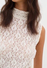 Kanten top in off-white, met een hoge halslijn, mouwloos ontwerp, ingewikkeld bloemenpatroon en gestructureerde stof.