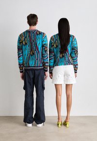 Knit sweater in vibrant blue, featuring multicolored patterns and ribbed cuffs. Pair of cargo pants and shorts, with green heels.