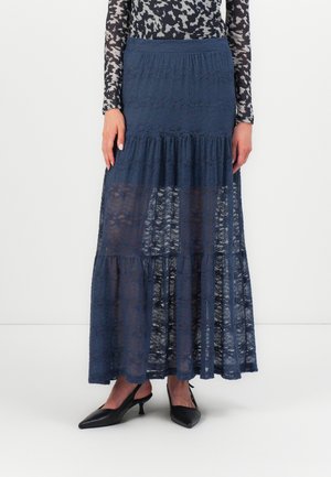 Femme portant une jupe maxi à volants en dentelle bleu marine avec des panneaux transparents, associée à des escarpins noirs à bout pointu et talon kitten, debout sur fond blanc.