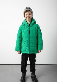 Dziecko noszące szary beret, zieloną kurtkę puchową, czarne spodnie cargo i czarne buty, stojące na szarej podłodze przy białej ścianie.