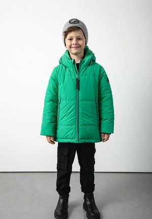 Dziecko noszące szary beret, zieloną kurtkę puchową, czarne spodnie cargo i czarne buty, stojące na szarej podłodze przy białej ścianie.