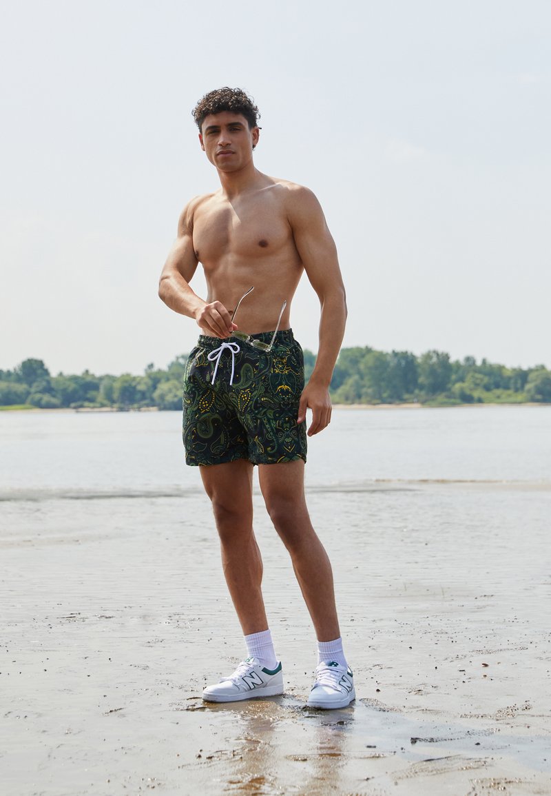 Dunkelgrüne Bade shorts mit einem Paisleymuster, weißem Kordelzug, kombiniert mit weißen Sneakers und Socken, an einem sandigen Strand.