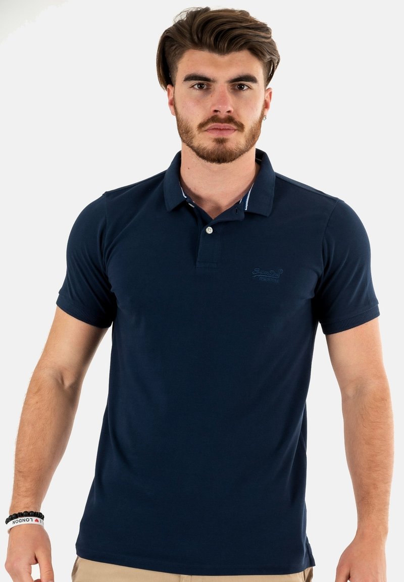 Polo shirt blu navy con colletto, maniche corte e logo sul petto. Presenta un sottile dettaglio a righe all'interno del colletto.