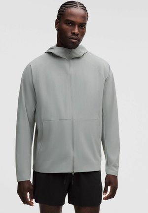Mand med flettet hår iført lyst gråt lynlåstrøje og sorte shorts, der står mod en ensfarvet baggrund.
