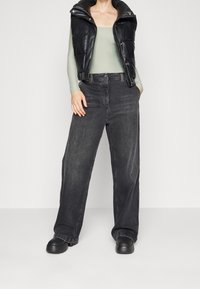 Osoba ubrana w czarną skórzaną kamizelkę puchową bez rękawów, na jasnoszarym longsleeve, w luźnych czarnych dżinsach i czarnych butach, stojąca z rękami w kieszeniach.