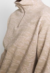 Beige välikerrospaita, jossa on neljänneksen mittainen vetoketjukaulus, hienovarainen valkoinen kuvio ja etuosassa kirjailtu "LEVI'S"-logo.