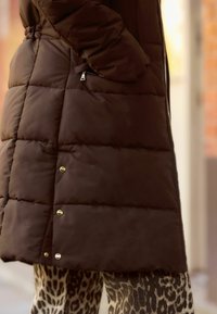 Doudoune marron avec un matelassage horizontal, poche zippée et boutons-pression au bas. Associée à un pantalon à imprimé léopard.