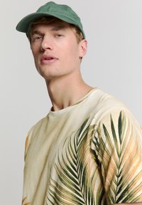 Groene pet gedragen met een kortebloes beige shirt met grote palmbladprints in groen en geel aan de voorkant. Neutrale achtergrond.