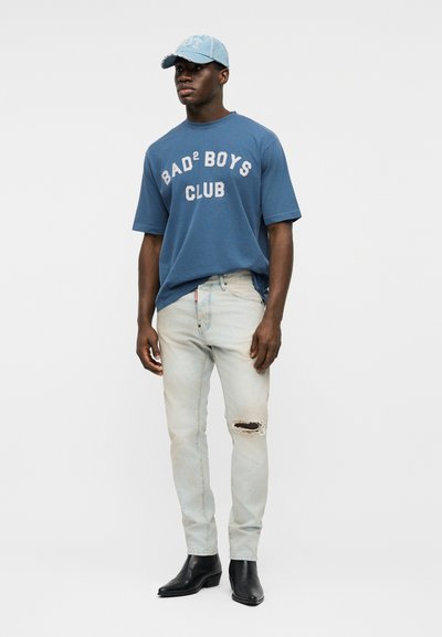 Νέος άνδρας που φοράει μπλε μπλούζα με το λογότυπο "BAD² BOYS CLUB", ανοιχτόχρωμα τζιν με σκισίματα, μαύρα μποτάκια και ανοιχτό μπλε καπέλο, στέκεται μπροστά σε απλό φόντο.