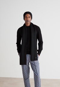 Schwarze Strickmütze und Schal kombiniert mit einem schwarzen Langarmshirt und gemusterten blauen Hosen, die geometrische Formen und Akzente aufweisen.