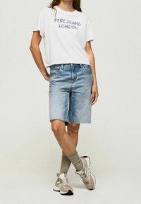 Camiseta blanca de manga corta con el texto "PEPE JEANS LONDON", combinada con pantalones cortos de mezclilla azul claro hasta la rodilla, calcetines grises y zapatillas multicolores.