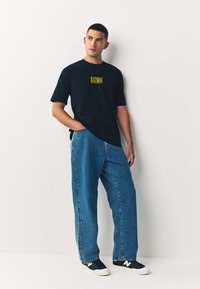 Camiseta negra con texto amarillo "BATMAN", combinada con pantalones vaqueros azul holgados y zapatos oscuros con detalles blancos.