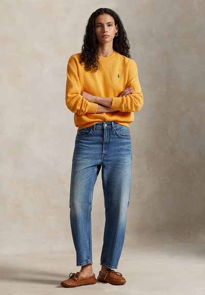 Żółta bluza z okrągłym dekoltem, niebieskie jeansy o prostych nogawkach, brązowe mokasyny. Kręcone włosy rozpuszczone, ręce skrzyżowane, neutralne tło.