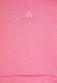 Felpa rosa realizzata in un tessuto morbido, con un piccolo logo Adidas in rilievo sul petto e un orlo arrotondato nella parte inferiore.