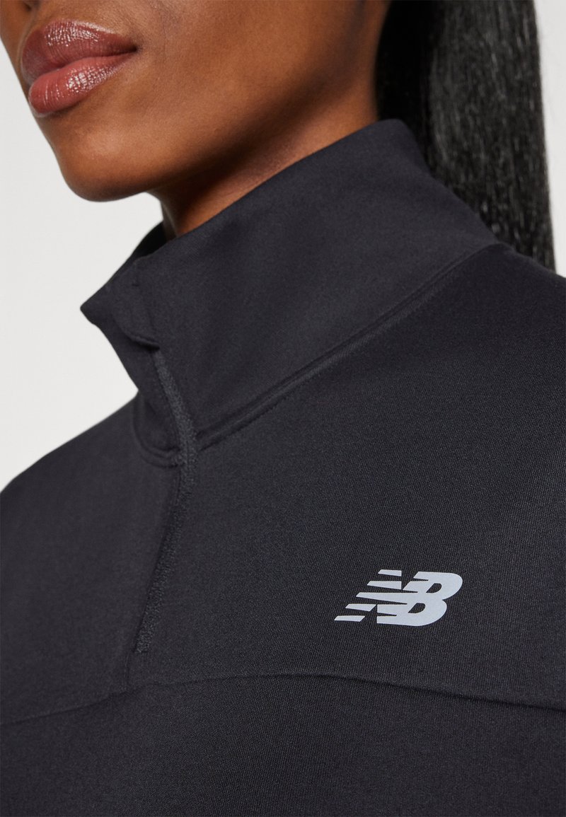 Top sportivo nero con colletto alto, dotato di chiusura a zip. Presenta un logo bianco di New Balance sul petto, tessuto di texture liscia.