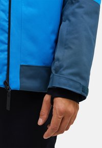 Wasserabweisende blaue Jacke mit dunkelblauem Ärmel, Reißverschlussverschluss und Klettverschluss am Ärmel. Verfügt über eine glatte Oberfläche und kontrastierende Farbpanele.