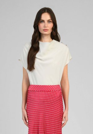 Femme aux longs cheveux bruns portant un haut blanc à manches courtes et une jupe rouge taille haute à pois blancs sur fond gris.