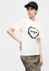 Γυναίκα που φοράει λευκό T-shirt Carhartt WIP με μαύρη καρδιά που μοιάζει να έχει βαφτεί με σπρέι, παντελόνι παραλλαγής και μαύρο καπέλο, στέκεται με το χέρι στη μέση.