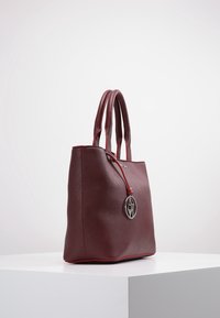 Vínově červená taška přes rameno s konstrukčním designem, hladkou texturou, červeným detailem podél okraje a stříbrným přívěskem s logem. Dvojité úchyty pro nošení.