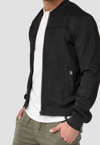 Veste bomber en daim noir avec fermeture éclair à l'avant, poignets et ourlet côtelés, et un logo discret sur la poche latérale. Portée sur une chemise blanche.