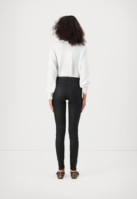 Jean noir taille haute avec une texture lisse, associé à un chemisier blanc à manches longues avec des poignets élastiques, vu de dos.
