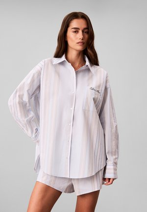 Vrouw met lang bruin haar die een oversized licht gestreept bloes met knopen en bijpassende korte broek draagt tegen een effen grijze achtergrond.