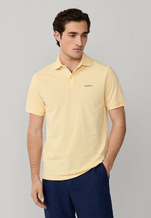 Jeune homme portant un polo jaune pâle avec le logo "Hackett" et un pantalon bleu marine, debout devant un fond uni gris clair.