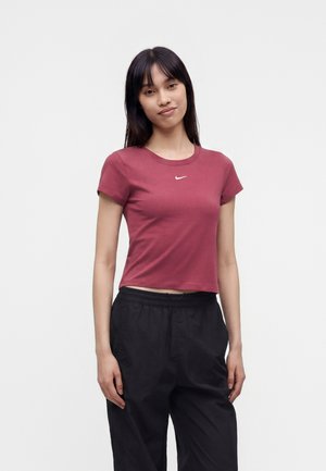 Giovane donna con capelli neri lisci, indossa un crop top bordeaux a maniche corte e pantaloni neri larghi, in piedi davanti a uno sfondo chiaro e uniforme.
