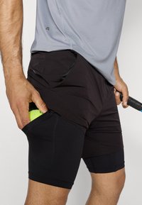 Muškarac u crnim kompresijskim kratkim hlačama ispod crnih teniskih hlača stavlja tenisku lopticu u džep na bedrima i drži teniski reket.