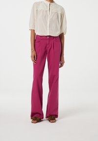 Pantalones de pierna ancha rosa con pliegues frontales, combinados con una blusa crema con detalles bordados y un cuello alto. Calzado con estampado de leopardo.