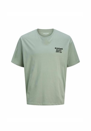 Camiseta de algodón verde claro con cuello redondo. Presenta un texto negro que dice "OLDWOOD DEEPLY ROOTED" en el lado izquierdo del pecho. Mangas cortas.