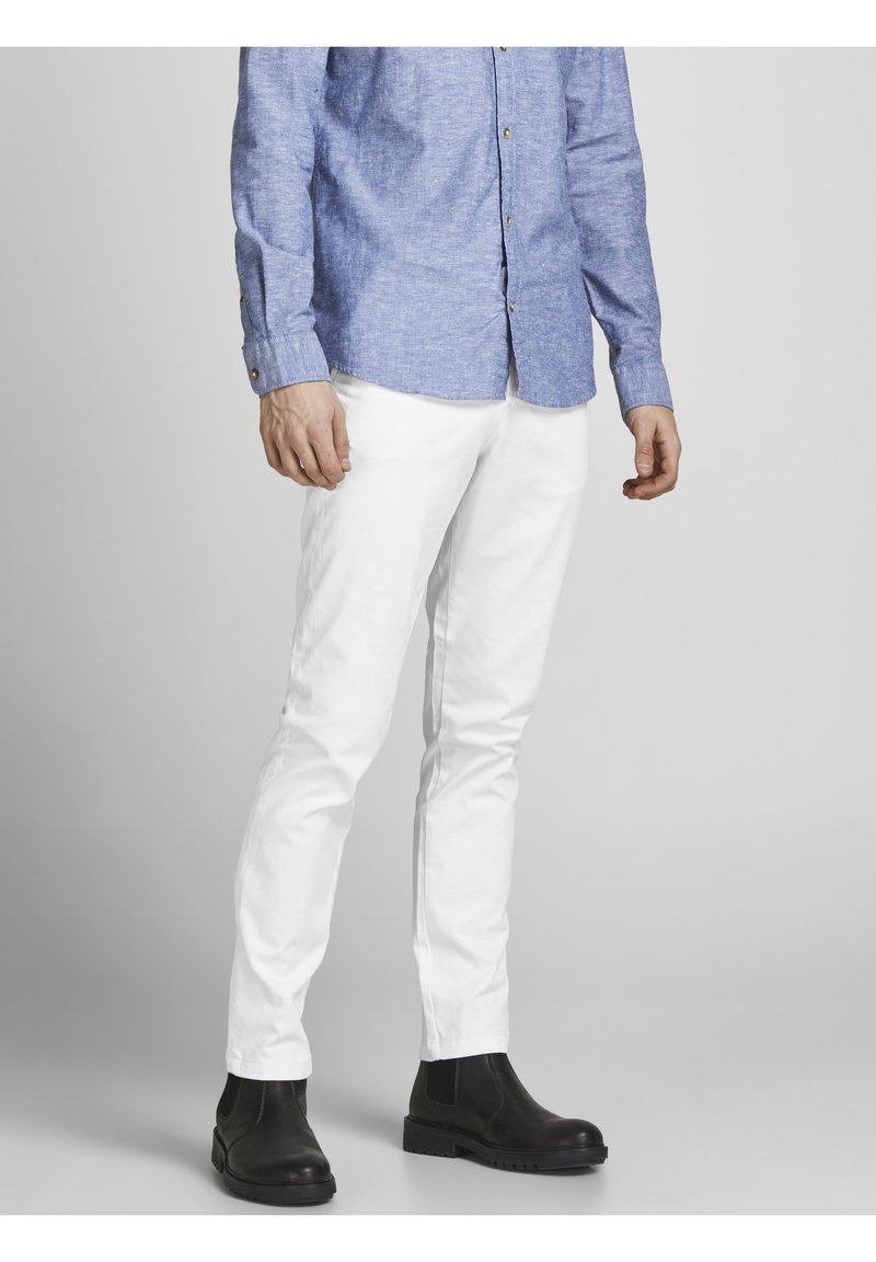 Homme portant un pantalon blanc, des bottes noires et une chemise bleue boutonnée, debout devant un fond gris uni.