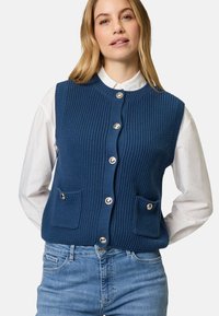 Dunkelblauer gestrickter Pullover mit Rippstruktur, rundem Ausschnitt, fünf großen Knöpfen und zwei vorderen aufgesetzten Taschen. Über einem weißen Hemd getragen.