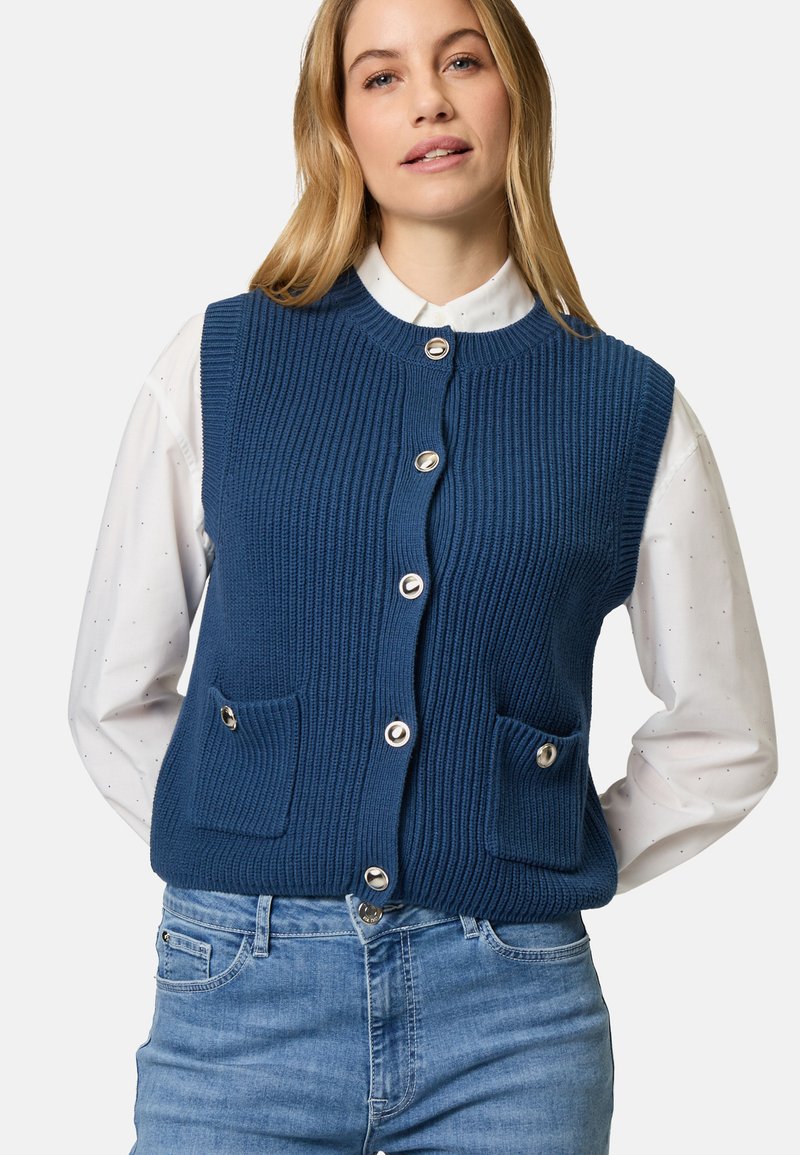 Dunkelblauer gestrickter Pullover mit Rippstruktur, rundem Ausschnitt, fünf großen Knöpfen und zwei vorderen aufgesetzten Taschen. Über einem weißen Hemd getragen.