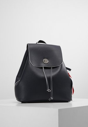 Sac à dos en cuir bleu marine avec un corps arrondi, une fermeture à rabat et des ferrures argentées. Il présente des surpiqûres blanches en accent et un panneau latéral coloré.