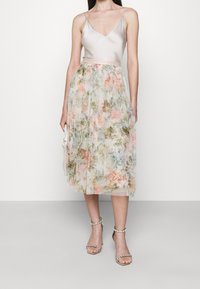 Kanten jurk met een licht satijnen lijfje en een zwierige, bloemmotief-plissérok in zachte pasteltinten. Wordt gedragen met sandalen met bandjes.