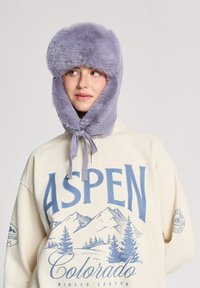 Jasnofioletowa czapka z futra syntetycznego z klapami na uszy, o miękkiej fakturze i dzianinowym sznurkiem na brodzie. Nosi ją osoba w beżowej bluzie z niebieskimi grafikami.