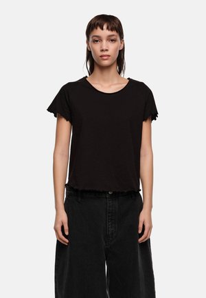 Joven con cabello oscuro y lacio, vistiendo una camiseta negra de manga corta y pantalones negros holgados, de pie contra un fondo liso.