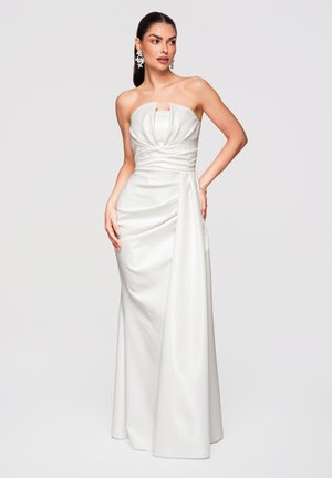 Femme portant une robe bustier en satin blanc avec corsage plissé et jupe drapée, posant avec la main sur la hanche devant un fond uni.