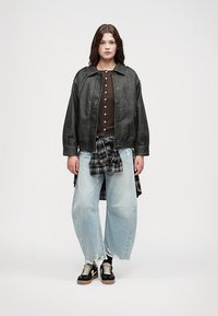 Casaco de pele preto, camisa de botão castanha, jeans de perna larga desgastados em azul claro, camisa de quadros atada à cintura, sapatilhas pretas com solas em tom bege.