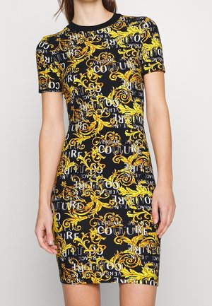 Kvinde iført en tætsiddende sort kjole med korte ærmer, dækket af gule barokmønstre og hvid Versace Couture tekst.