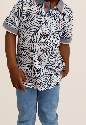 Kind trägt ein weißes Kurzarm-Poloshirt mit blauen, schwarzen und roten Blattmustern, blauem Kragen und Ärmelsäumen, kombiniert mit blauen Jeans.