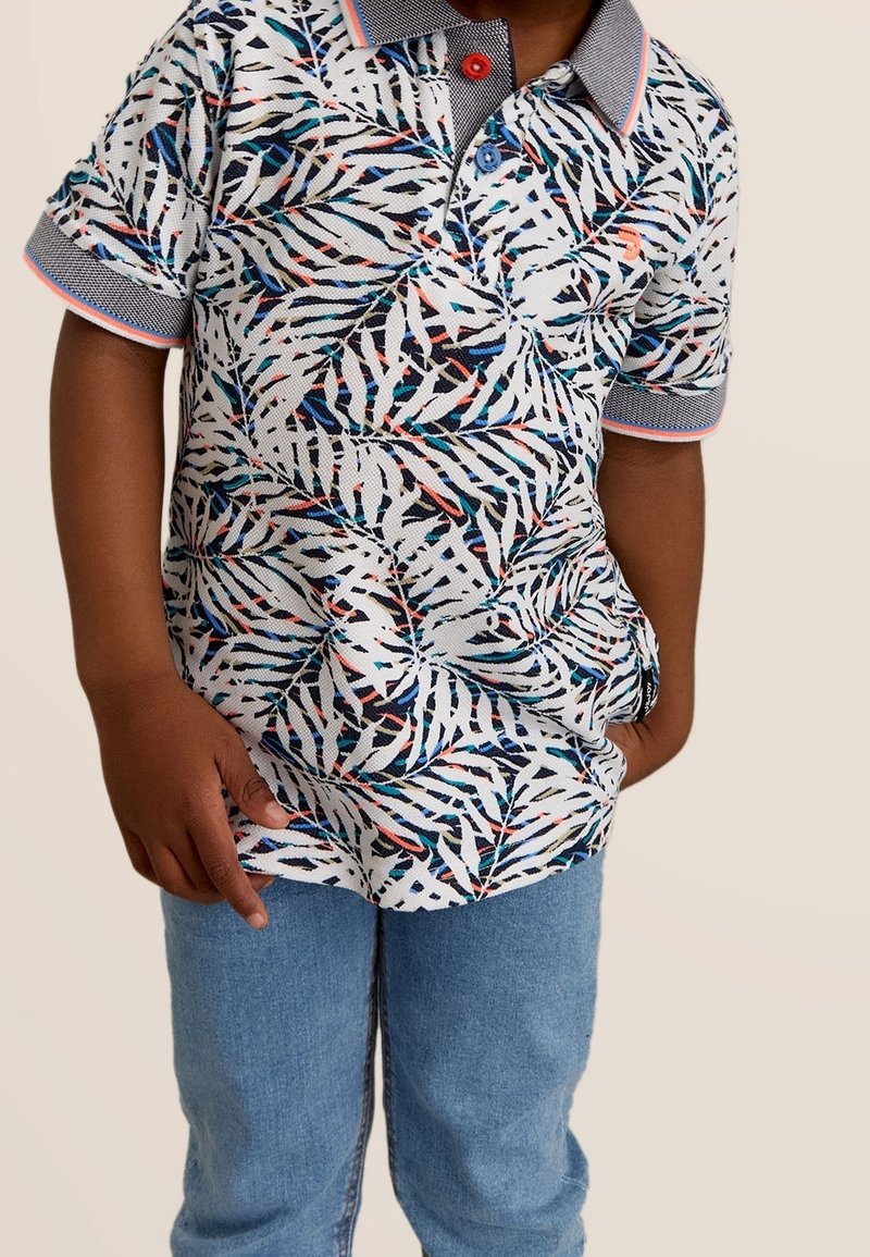 Enfant portant un polo blanc à manches courtes avec des motifs de feuilles bleues, noires et rouges, col et bords de manches bleus, associé à un jean bleu.