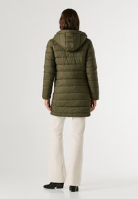 Olīvzaļš pūkains mētelis ar kapuci, kas piedāvā horizontālu šuvju rakstu, vidējo garumu un taisnu piegriezumu. Apvienots ar krēmkrāsas svārku biksēm.