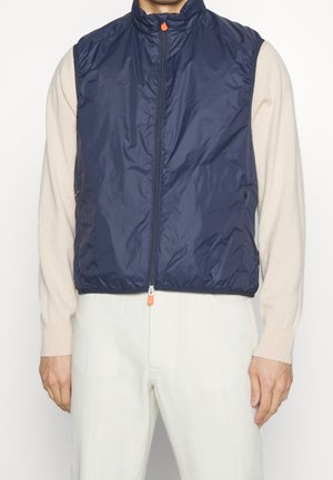 Man wearing a navy blue sleeveless zip-up vest over a cream long-sleeve sweater and light-colored pants against a plain background.