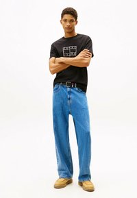 Zwarte t-shirt met een wit logo, lichtblauwe baggy jeans en tan suede schoenen. De outfit heeft een zwarte riem met een kleuraccent.